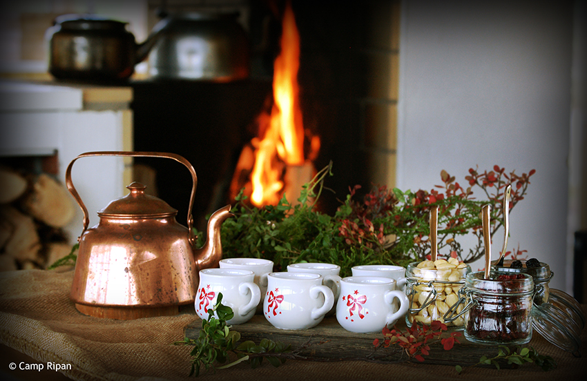 Christmas decorations around a coffee pot, cups and gluhwein toppings and a fire in the background